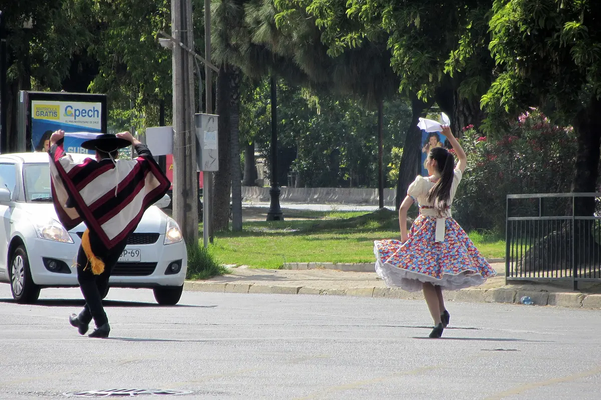A dancer in a poncho and hat and a dancer in a flowered full skirt, both with handkerchiefs raised, dancing the cueca on a Santiago street